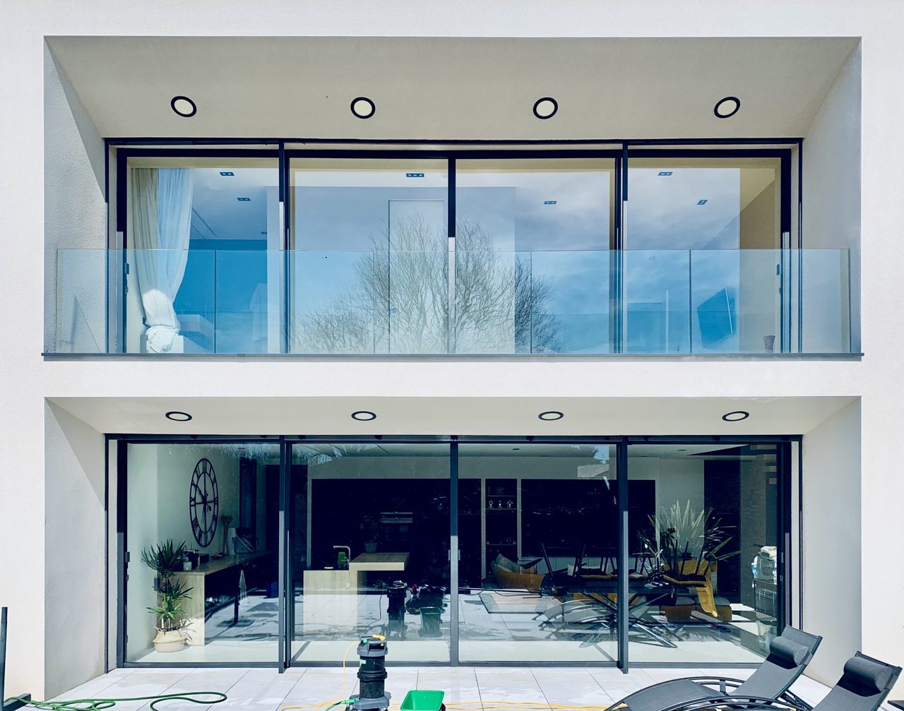 Logement contemporain à grandes baies vitrées et balcon en verre, avec le matériel de nettoyage posé sur la terrasse.