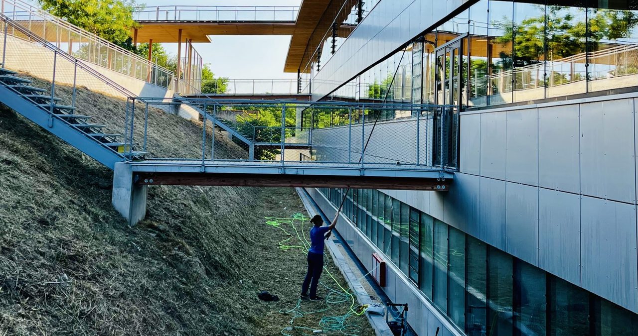 Technicien Clean-Code nettoyant les vitrages d’un collège avec une perche télescopique, depuis le sol, sous une passerelle.
