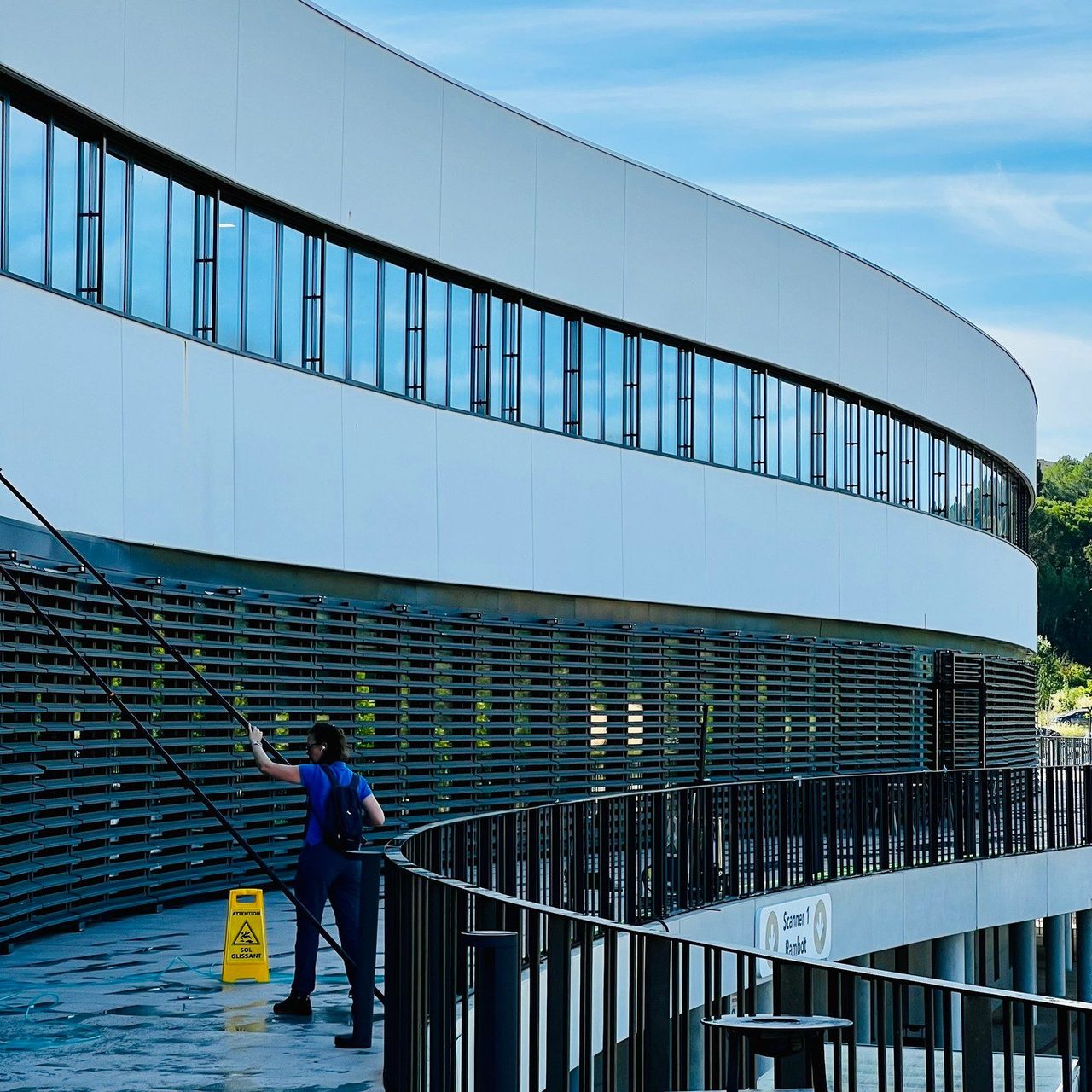 Technicien Clean-Code, vu de dos, nettoyant à la perche la façade vitrée courbe de l’hôpital privé HPP. Un panneau sol glissant est posé au sol.