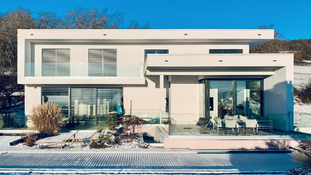 Maison contemporaine blanche avec de grandes baies vitrées, un garde-corps en verre et une terrasse, sous un ciel d’hiver.